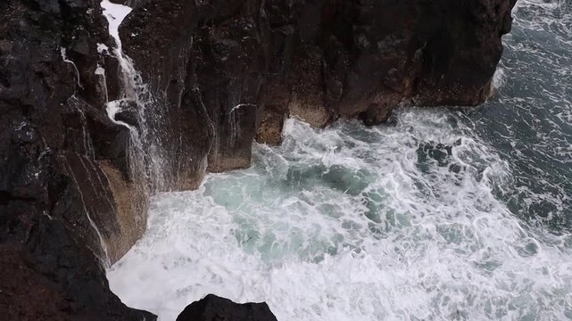 A beautiful view of a rocky volcanic basin with a storm wave crashing against it, creating foamy spray, on the northern coast of an Atlantic Ocean island, viewed from above, close-up, and in real moti