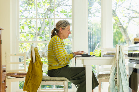adult woman sitting at table in light filled room using mobile phone and typing on laptop on table