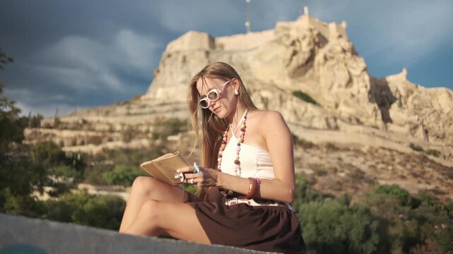 Stylish young woman writing in her journal while sitting outdoors with a historic castle in the background during sunset