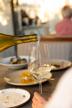 waiter or sommelier pouring white wine into a glass an a restaurant with outdoor patio and pizza and pasta on the table, depth of field with other tables