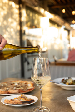 waiter or sommelier pouring white wine into a glass an a restaurant with outdoor patio and pizza and pasta on the table, depth of field with other tables
