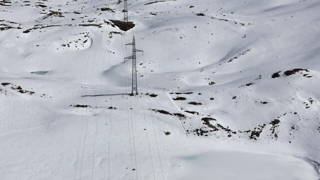 power pylons cross broad snowfield pale meltwater pond nestles below scattered rock patches faint road traces distant edge julier pass alpine crossing graubunden switzerland high altitude