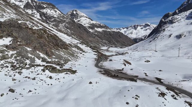 paved road winds through snowbound valley toward jagged peaks single car travels route clear blue sky overhead julier pass mountain crossing graubunden switzerland high altitude