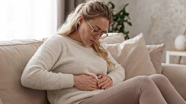 A mature blonde woman sits on a couch and suffers from sharp stomach pain in a bright cozy living room.