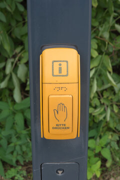 Close-up of a yellow public transit or information button with braille and German text bitte dr&uuml;cken on a post against a green foliage background.