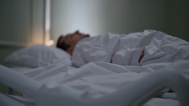 Sick patient sleeps calmly on adjustable clinic bed in dark room. Adult male rests peacefully beneath white bedsheets, showing somber healthcare situation during nighttime observation.