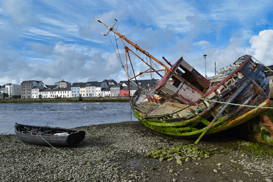 Abandoned wooden fishing boat wreck on the shore with colorful Claddagh houses in Galway Ireland