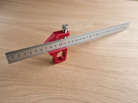 Red Marking Gauge With Steel Ruler on - A red aluminum marking gauge with a stainless steel 30cm ruler placed diagonally on a light wood surface.