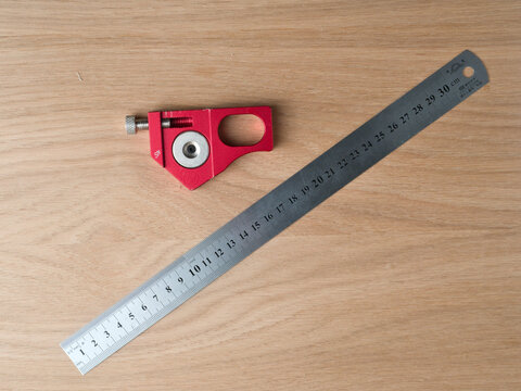 Red Marking Gauge With Steel Ruler on - A red aluminum marking gauge with a stainless steel 30cm ruler placed diagonally on a light wood surface.