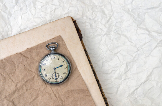 Vintage pocket watch rests on an old book and wrinkled paper. The still life evokes memory and time. Copy space supports history, nostalgia, and design.