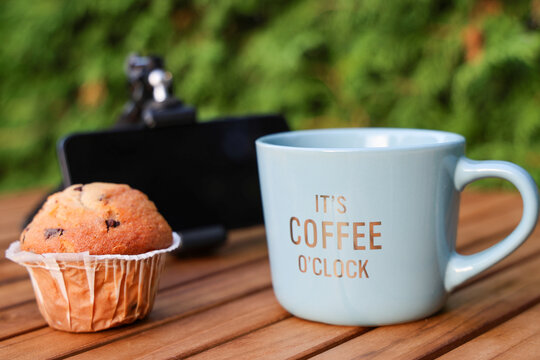 Blue coffee mug and chocolate muffin on wooden table outdoors with blurred smartphone on mini tripod in background, cozy morning break concept.