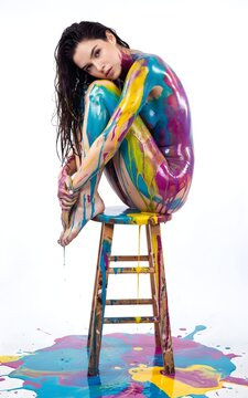 Modelo femenina con el cuerpo cubierto de pintura colorida sentada en un taburete de madera sobre fondo blanco