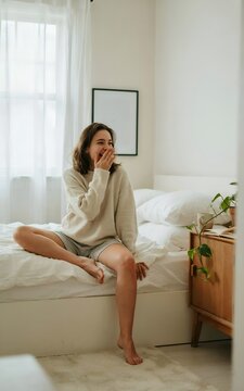 Joven mujer relajada bostezando alegremente sentada al borde de la cama en un dormitorio luminoso por la ma&ntilde;ana