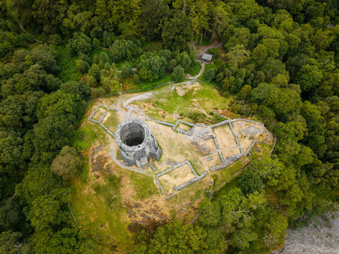 Medieval castle ruins with circular stone keep on grassy hilltop, ancient foundations surrounded by lush green forest, overhead drone shot