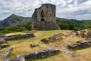 Wide shot of historic stone castle ruins with circular tower keep, ancient fortifications on rugged grassy hill with mountains behind. © whitcomberd