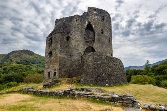 Low angle view of ancient ruined stone keep on grassy hill, dry stone wall in foreground under cloudy sky, historic fortification in remote landscape