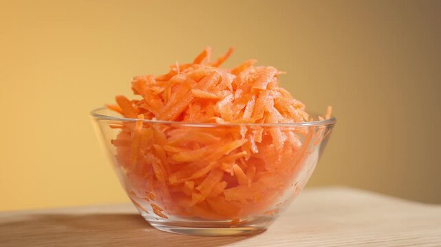 Freshly grated carrots in salad glass bowl rotate against orange background, closeup of vegetables, preparing healthy dieting vegan meal