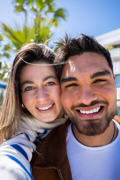 Vertical. Close portrait of a happy couple smiling directly at camera while standing outdoors in a bright modern residential area with palm trees and blue sky.
