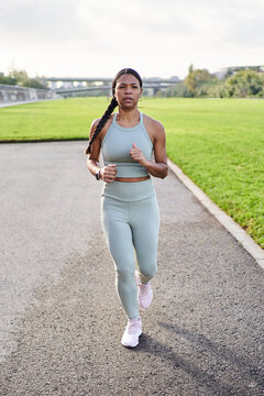Vertical. Full body african millennial woman jogging outdoors in park on sunny day, wearing sportswear and smartwatch. Expression shows energy, discipline and determination in active lifestyle.