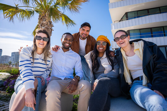 Multicultural group portrait of friends seated together in a bright residential district with palm trees, modern buildings and warm sunny light.