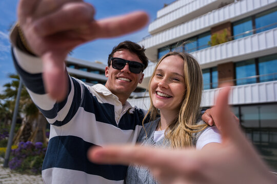 Happy young couple smile and reach their hands toward the camera in a playful wide-angle portrait outdoors in a sunny modern urban setting.
