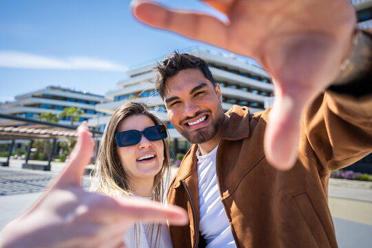 Young man and woman smile at camera while making a hand frame outdoors in a bright residential district with modern architecture and clear sky.