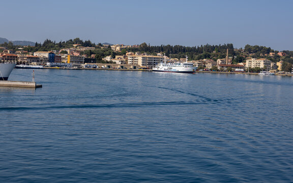 passenger ferry docked at corfu island harbor with traditional coastal town, lush green hills, and calm blue ionian sea under a clear sunny sky in greece.