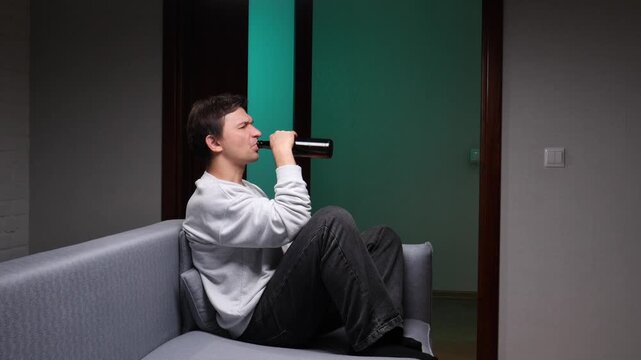 Young man sitting alone on a sofa, drinking alcohol directly from a bottle, depicting themes of depression, stress, loneliness, and alcoholism in a dim lit room