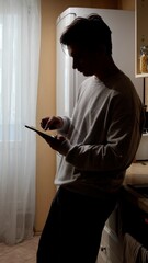 Young man standing in a home kitchen, leaning on the counter, engaging with a digital tablet and browsing content, representing modern lifestyle and connectivity © Aliaksandr