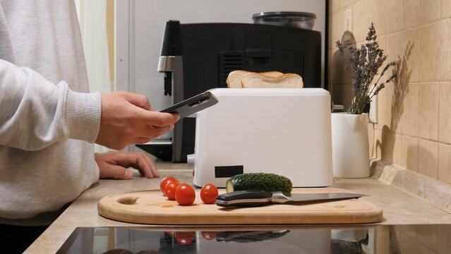 Person using smartphone to remotely control a white smart toaster with bread slices on a modern kitchen counter, preparing a convenient, connected breakfast routine at home