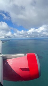 Airplane wing with red engine flying over blue ocean and clouds, showcasing a scenic aerial view of the coastline and water surface. 4K vertical footage