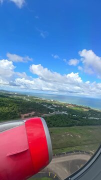 Airplane wing and engine view over coastal landscape with blue sky and clouds during descent towards tropical destination. 4K vertical footage