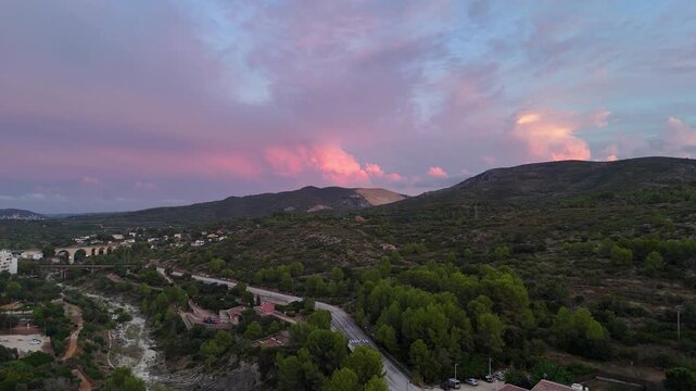 Aerial view of Gata de Gorgos, Alicante, Spain, under a dramatic pink and purple sunset sky, with a stone viaduct, a dry riverbed, pine trees and a winding road through the small town