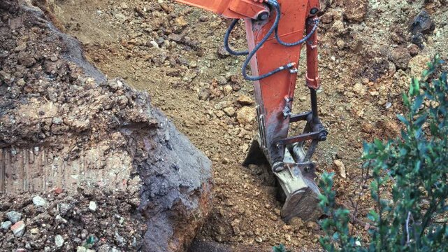 Close-up shot of an orange backhoe excavator bucket digging into and scooping up rocky soil and dirt at a construction site foundation base.