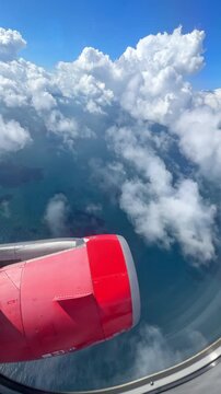 Airplane wing with red engine above clouds and ocean, showcasing a clear blue sky and scattered white clouds in the background. 4K vertical footage