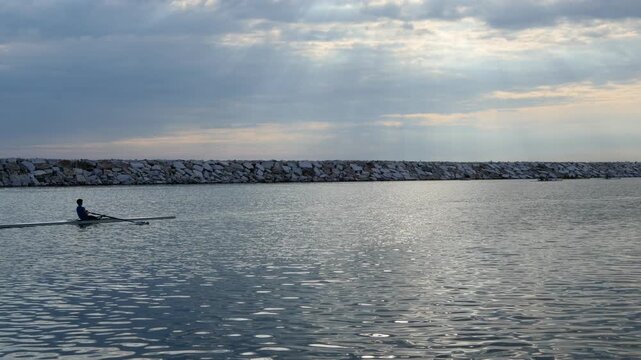 A single rower sculls across calm coastal waters parallel to a stone breakwater under a dramatic cloudy sky with sunbeams in Kucukyali, Istanbul.