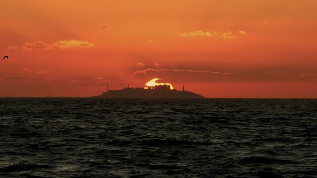 Telephoto shot of Yassiada island silhouetted against a vibrant orange sunset in the Sea of Marmara, with the sun setting directly behind the landmass and a bird flying in the dramatic sky. Istanbul, 