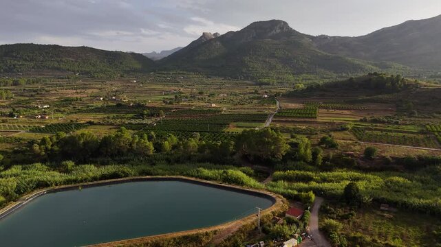Aerial drone footage of a patchwork farmland valley, olive groves and a circular irrigation reservoir near Alcalali, Marina Alta, Alicante, Spain, with a winding stream and mountain ridges