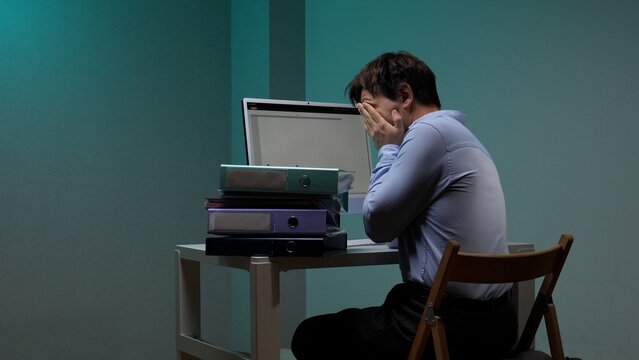 Overwhelmed businessman at a dark office desk, hands covering his face amid a laptop and towering binders, conveying burnout, exhaustion and deadline driven despair