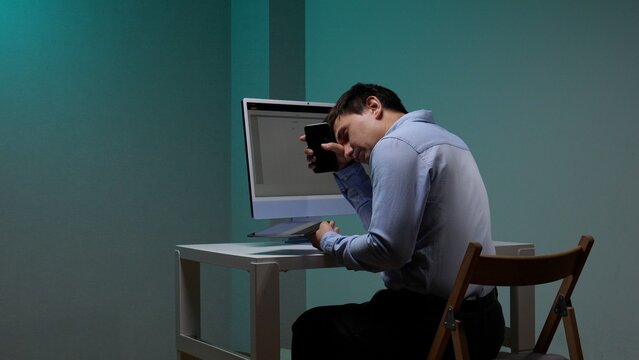 Young man experiencing burnout and fatigue, holding a smartphone to his face while sitting at a desk in front of a computer, highlighting the stress of overworking