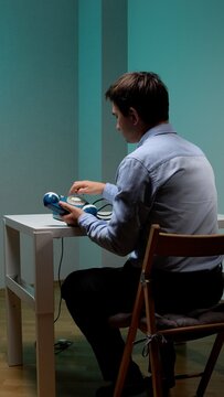 Young man sitting at a simple white desk, dialing a blue vintage rotary telephone, engaging in an old fashioned communication method in a minimalist room