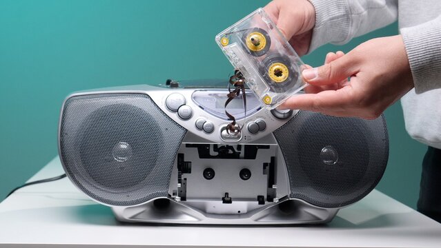 Human hands holding a damaged audio cassette tape with its magnetic ribbon tangled and pulled out, standing over an open retro boombox, symbolizing obsolescence and past technology issues