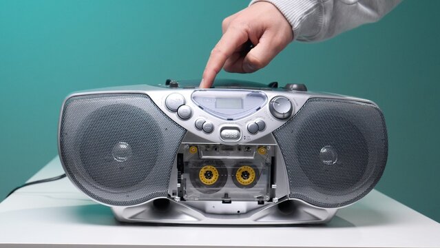 Hand pressing a control button on a vintage silver boombox with an audio cassette tape loaded, highlighting the classic music playback technology and nostalgic electronics on a white surface