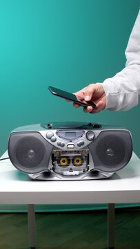 Hand holding a modern smartphone above a vintage portable boombox with an open cassette player, illustrating the evolution of music playback technology and generational changes in audio devices