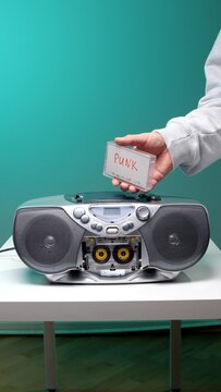 Hand holding a transparent cassette tape with punk written on it, ready to insert into an open, silver boombox on a white table, representing retro audio technology and nostalgia