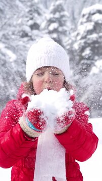 Child is having fun outdoors in winter forest. A girl blows away the snow in a nature park against a snow covered trees background. Slow motion. Vertical video