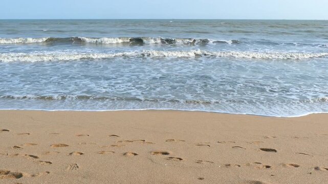 Handheld shot of beach shoreline with rolling sea waves and footprints on golden sand
