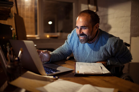 Smiling wheelchair user working late on laptop at home office