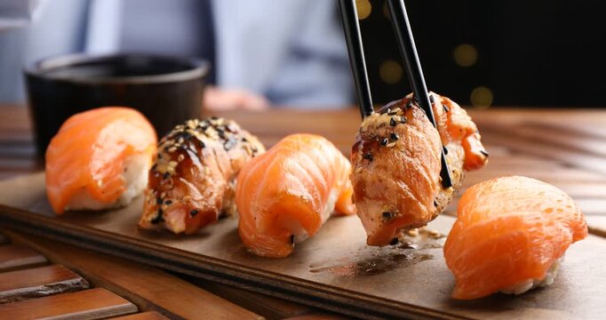 Woman taking delicious nigiri sushi with chopsticks from wooden table, closeup