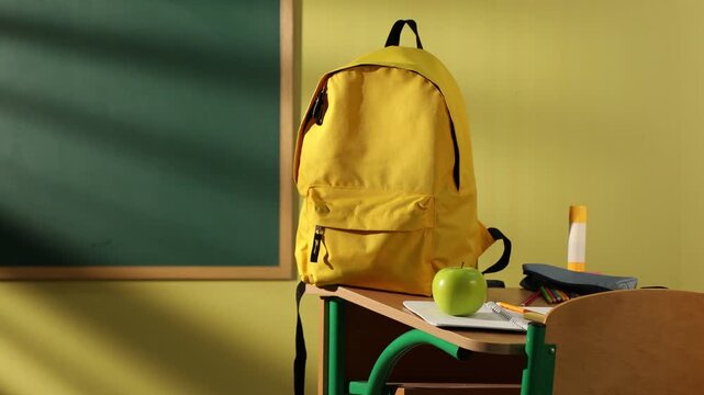 Stylish backpack with different stationery, glasses and apple on wooden desk indoors, camera moving left